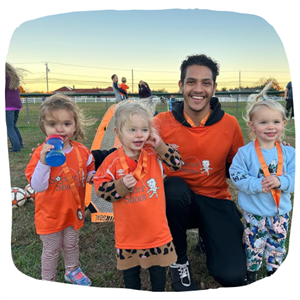 Soccer Coach in bright orange shirt with 3 small children proudly displaying their medals.